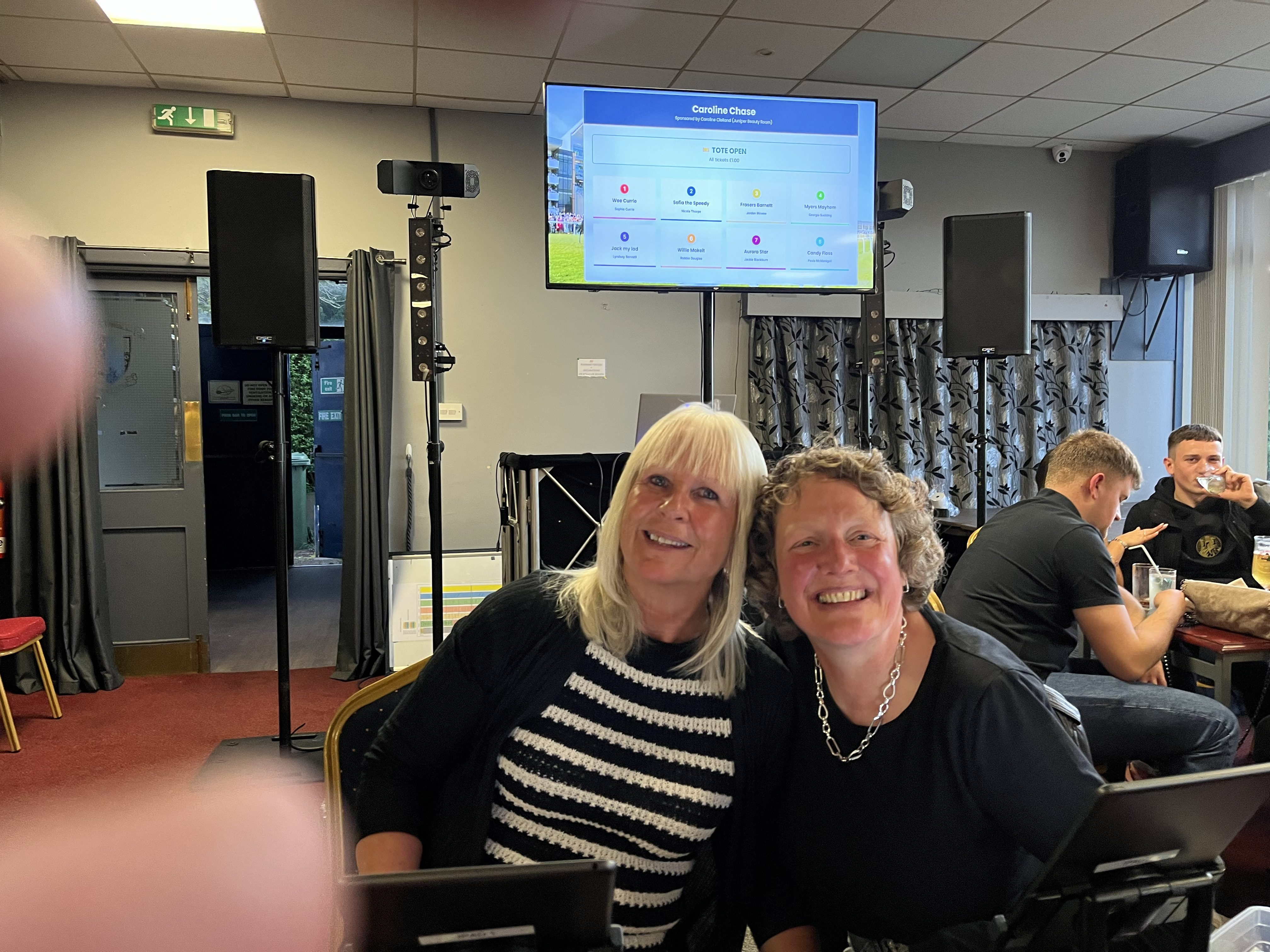 Two guests smiling at the Premier Disco race night tote board showing Caroline Chase race at Sighthill Bowling Club