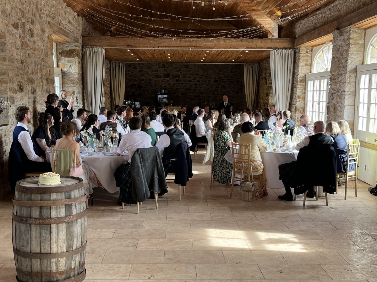 Wedding reception speeches at Colstoun House, East Lothian — guests seated at round tables in stone barn with string lights and whisky barrel cake stand