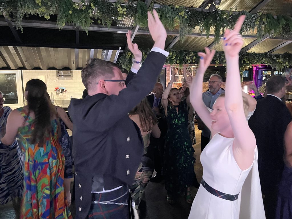 People at a lively party dancing with hands raised, a woman in a white dress in foreground, others in colorful outfits under hanging greenery.