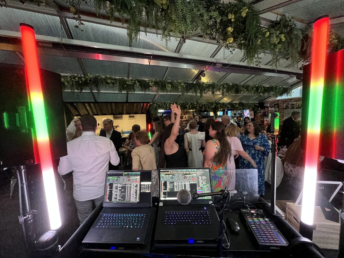DJ booth at Stewart Brewing wedding reception with light towers and laptops, guests visible on the dancefloor behind