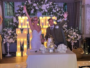 Bride and groom cut their white wedding cake, hands joined, with glowing ‘MR & MRS’ behind and white flowers.